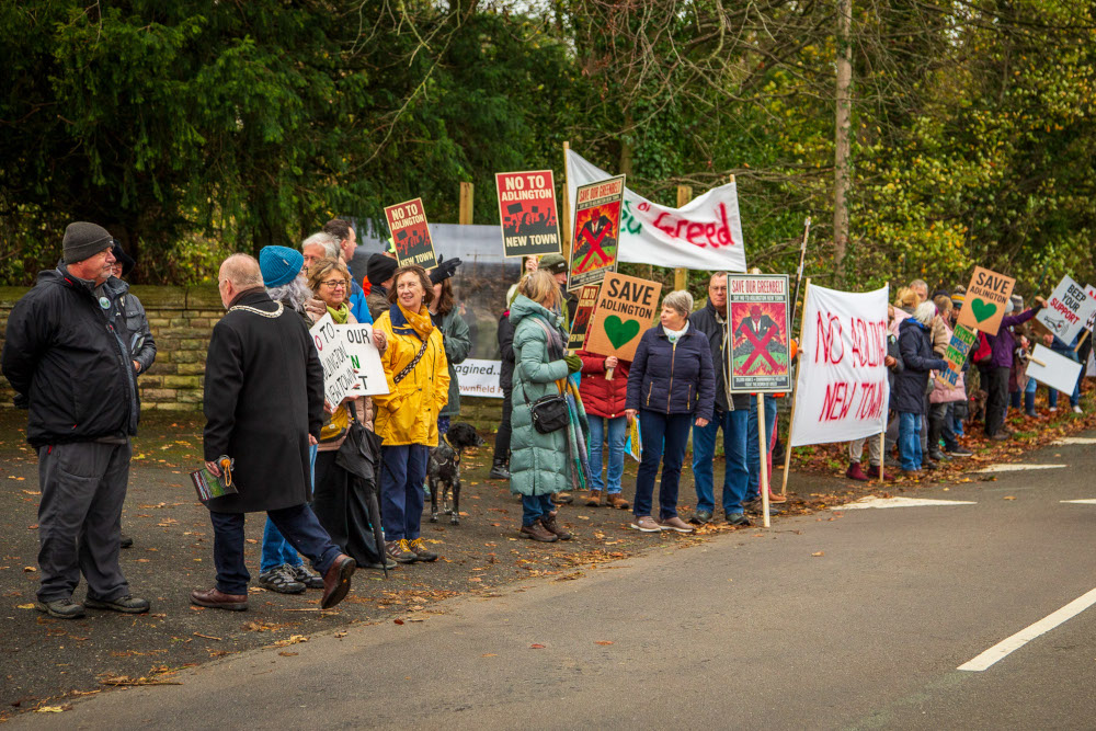Protesters on the side of the road outside Adlington Hall holding a variety of banners and placards