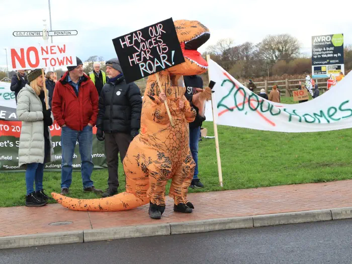 Dinosaur on roundabout in opposition to Adlington New Town holding a placard Hear Our Voice Hear Us Roar!